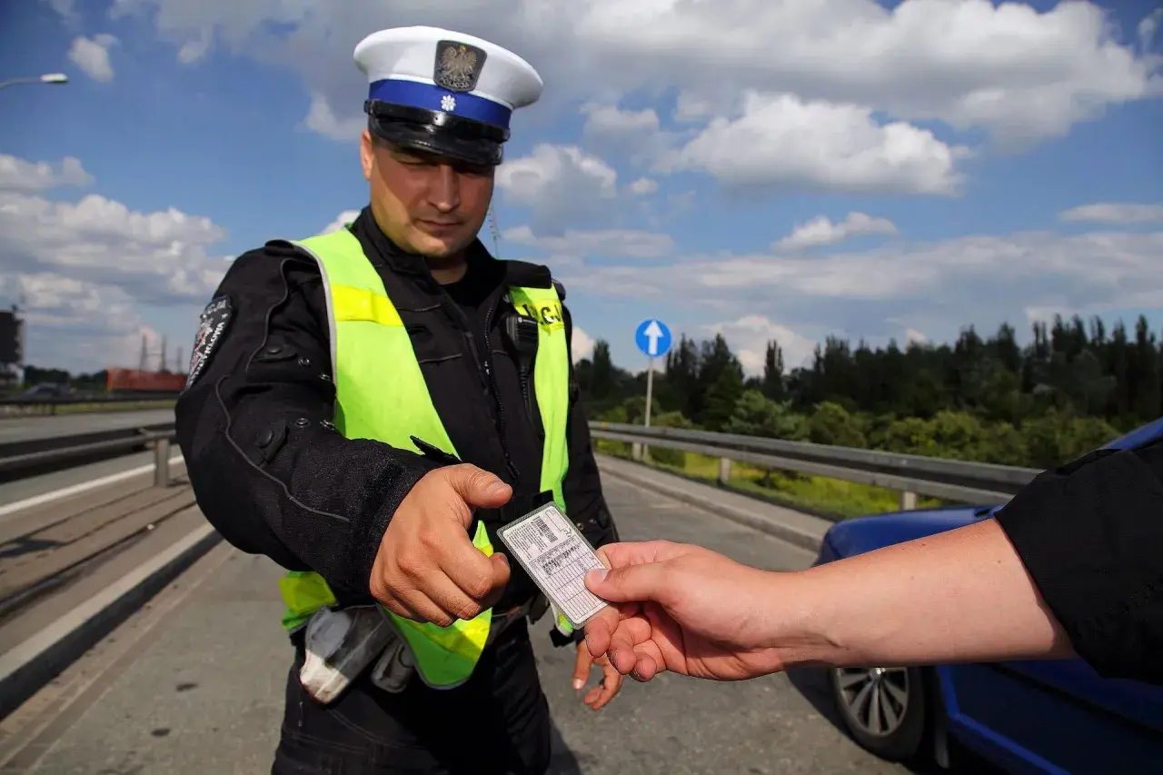 Policjant w kamizelce odblaskowej odbiera dokument. Czy trzeba mieć przy sobie prawo jazdy? Tak, podczas kontroli drogowej.
