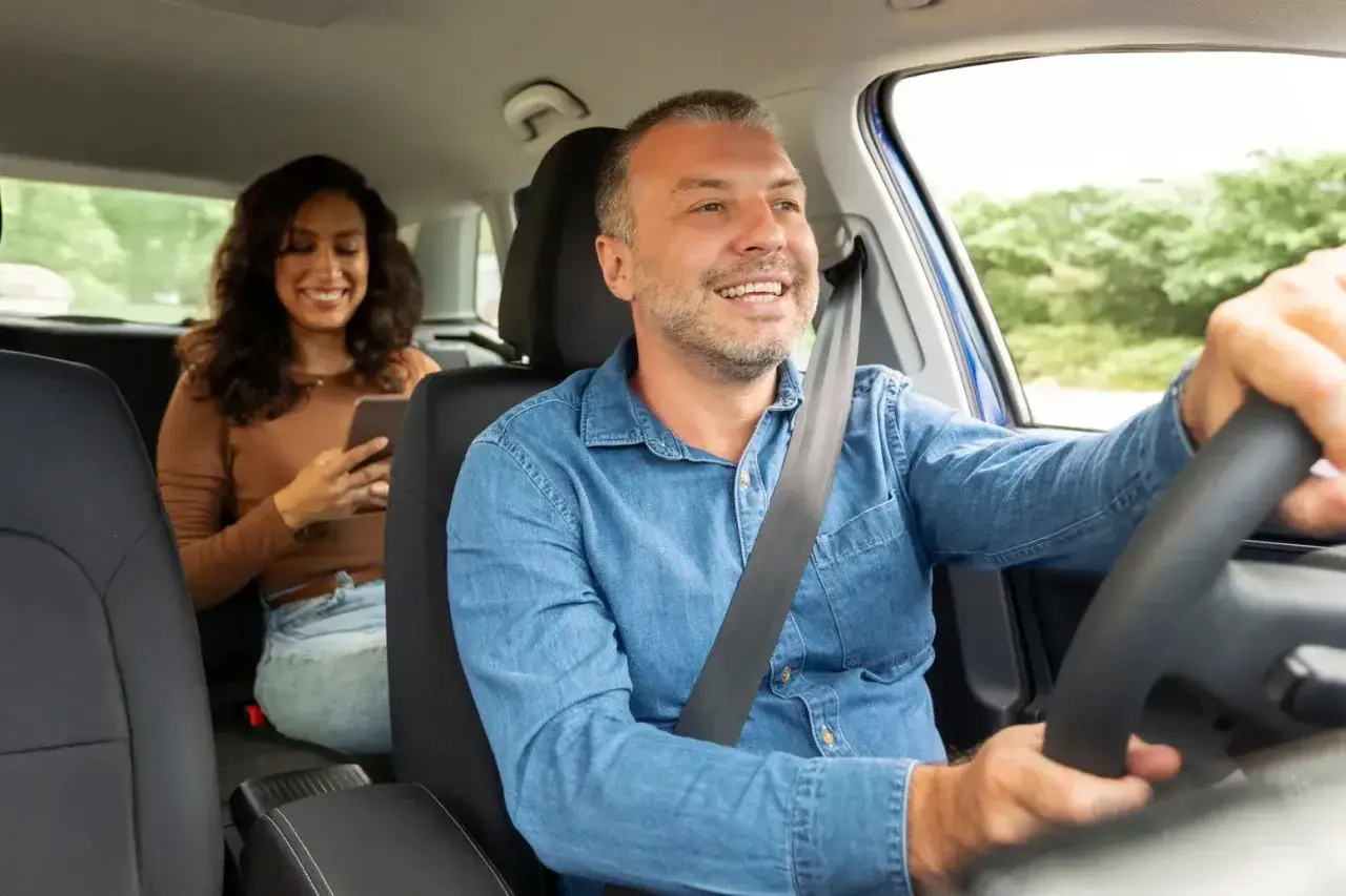 Uśmiechnięty kierowca taxi prowadzi samochód, pasażerka z tyłu korzysta z telefonu.