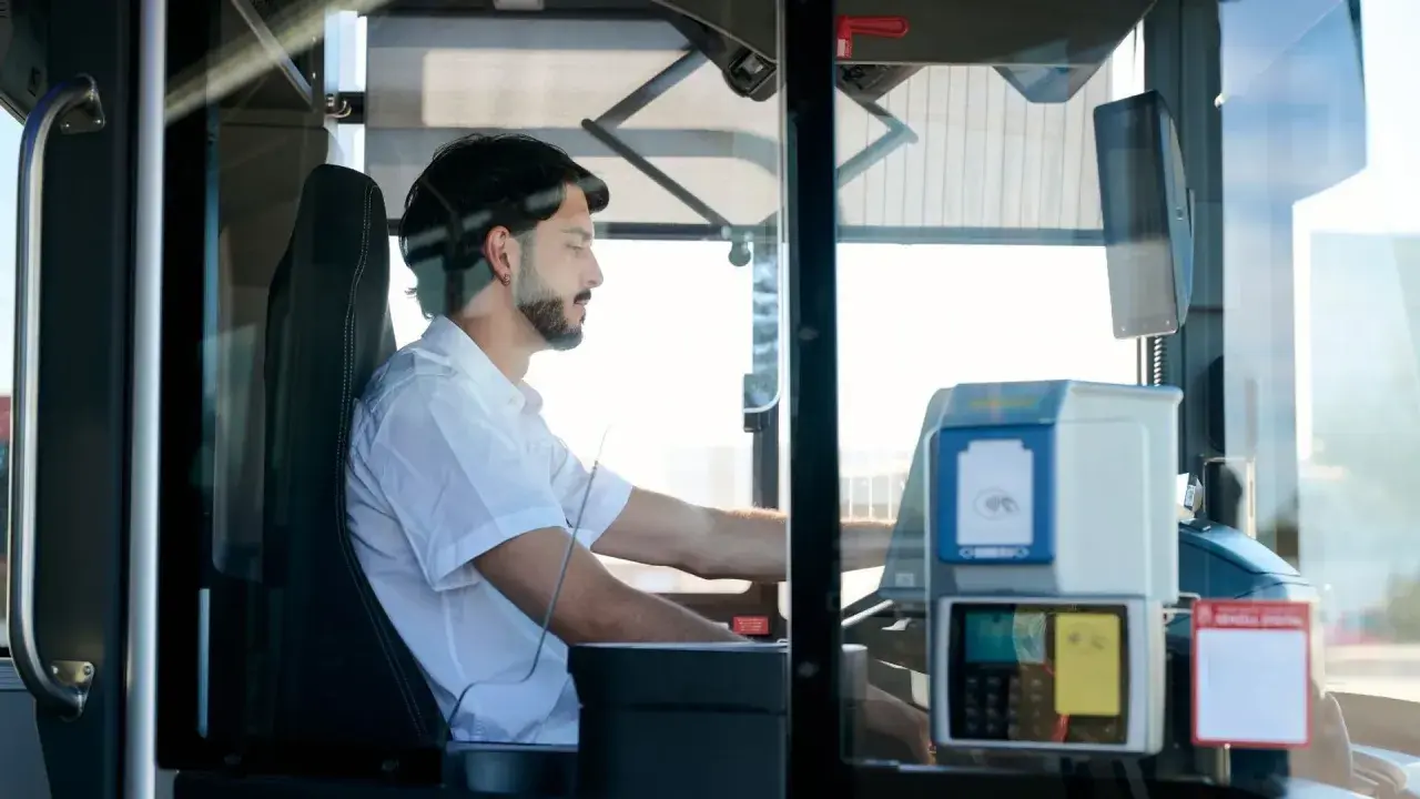 Mężczyzna za kierownicą autobusu, zastanawiający się, ile zarabia kierowca autobusu. Widoczny biletomat i siedzenie kierowcy.