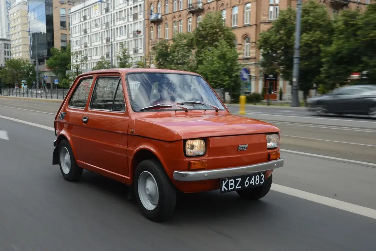 Pomarańczowy Fiat 126p jedzie ulicą. Jego pojemność baku to 25 litrów, co pozwala na długie podróże.