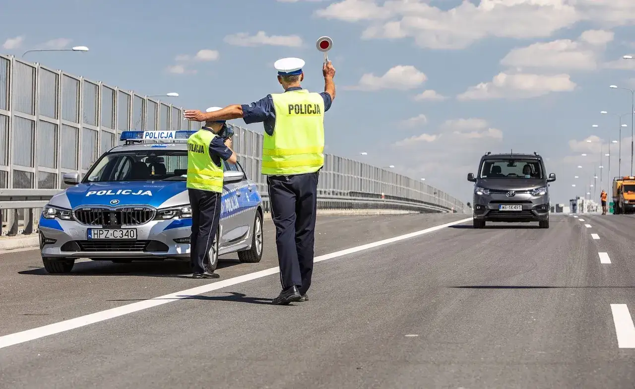 Policjanci na drodze zatrzymują pojazd. Jeden z nich podnosi znak stop, sygnalizując zatrzymanie prawa jazdy.