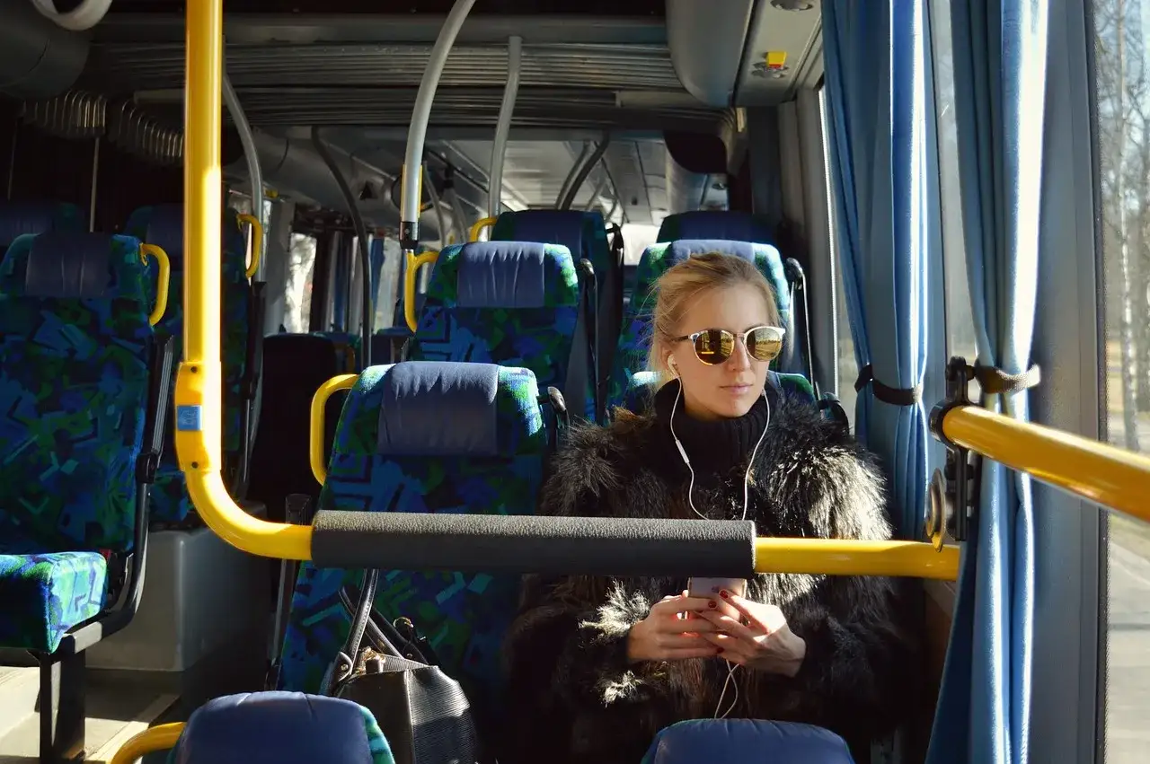 Kobieta w futrzanej kurtce i okularach przeciwsłonecznych słucha muzyki w autobusie, zastanawiając się, ile zarabia kierowca autobusu.