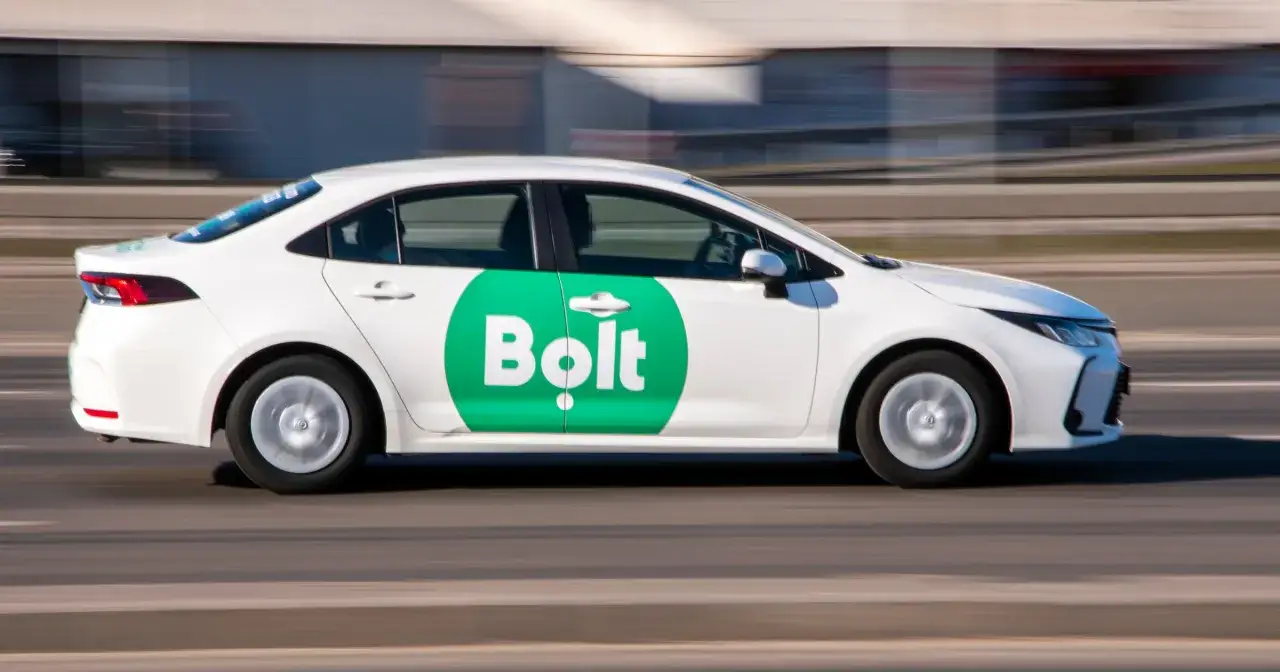 Biały samochód z zielonym logo Bolt na drzwiach jedzie po drodze. Chcesz wiedzieć, jak zostać kierowcą Bolta?