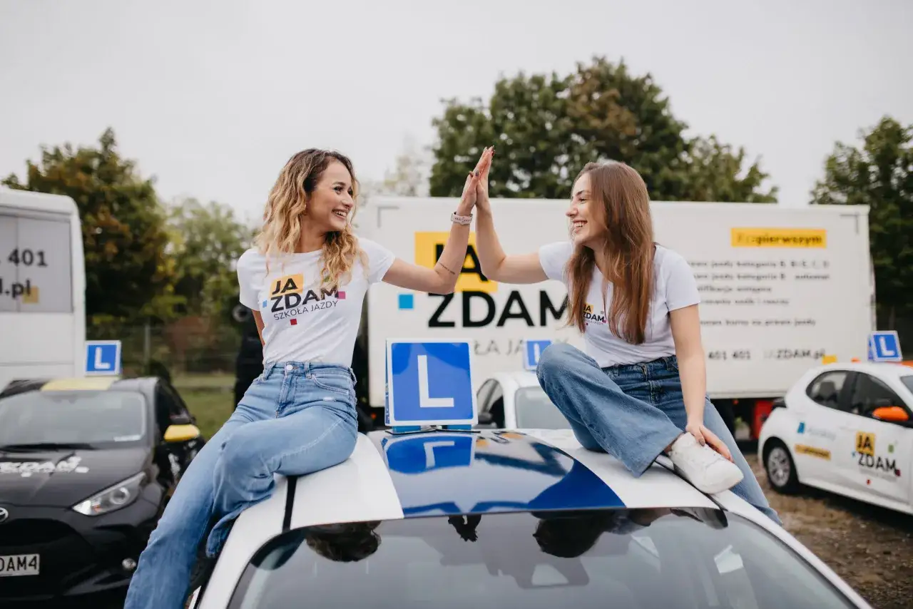 Dwie uśmiechnięte dziewczyny świętują sukces w szkole jazdy. Czy to oznacza, że zdobyły prawo jazdy na samochód jaka kategoria?