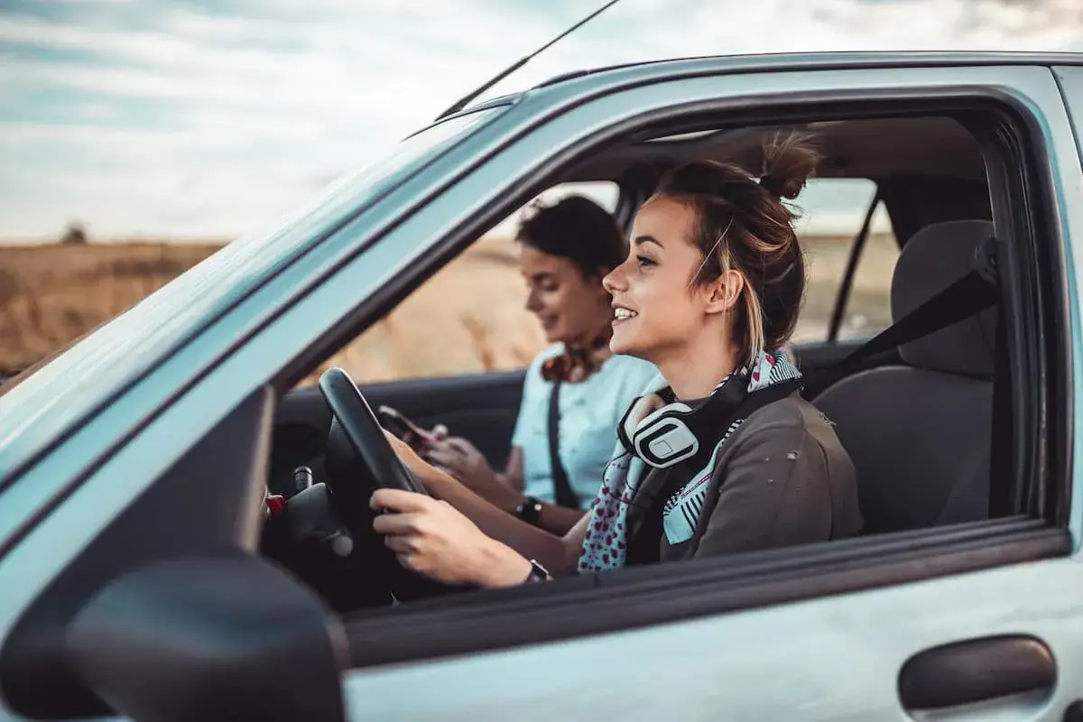 Młodociany kierowca, ile lat ma? Uśmiechnięta dziewczyna z słuchawkami na szyi prowadzi auto, pasażerka obok patrzy w telefon.