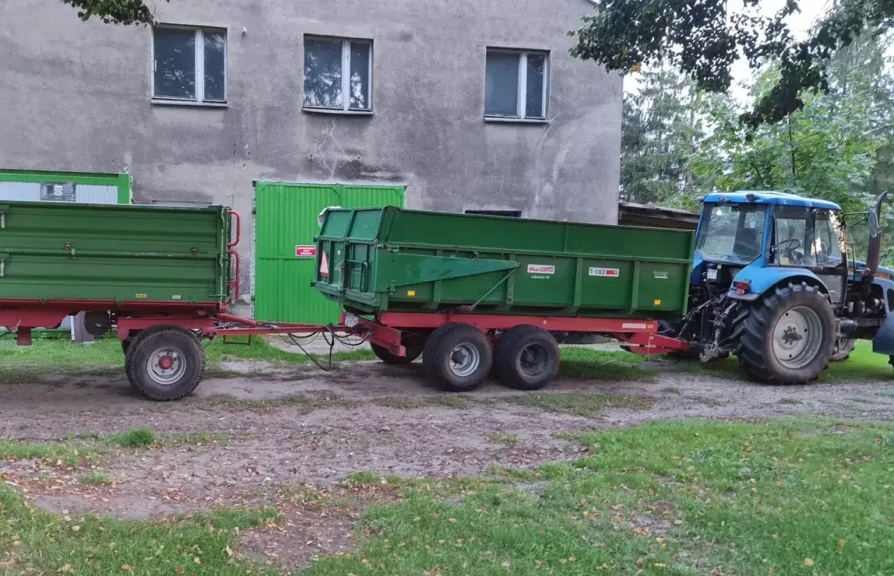Niebieski traktor z dwoma zielonymi przyczepami. Zastanawiasz się, czy na traktor trzeba mieć prawo jazdy?