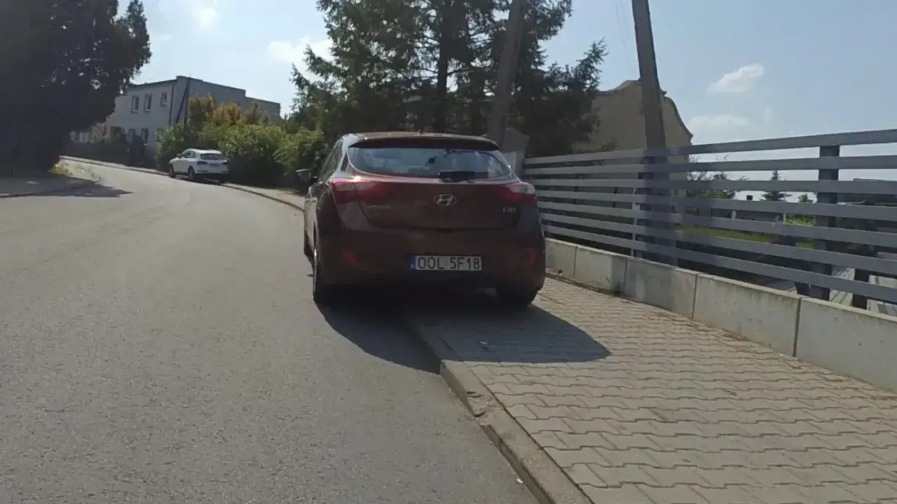 Czerwony Hyundai z tablicą rejestracyjną OOL 5F18 stoi na poboczu drogi. W tle budynek i drzewa.