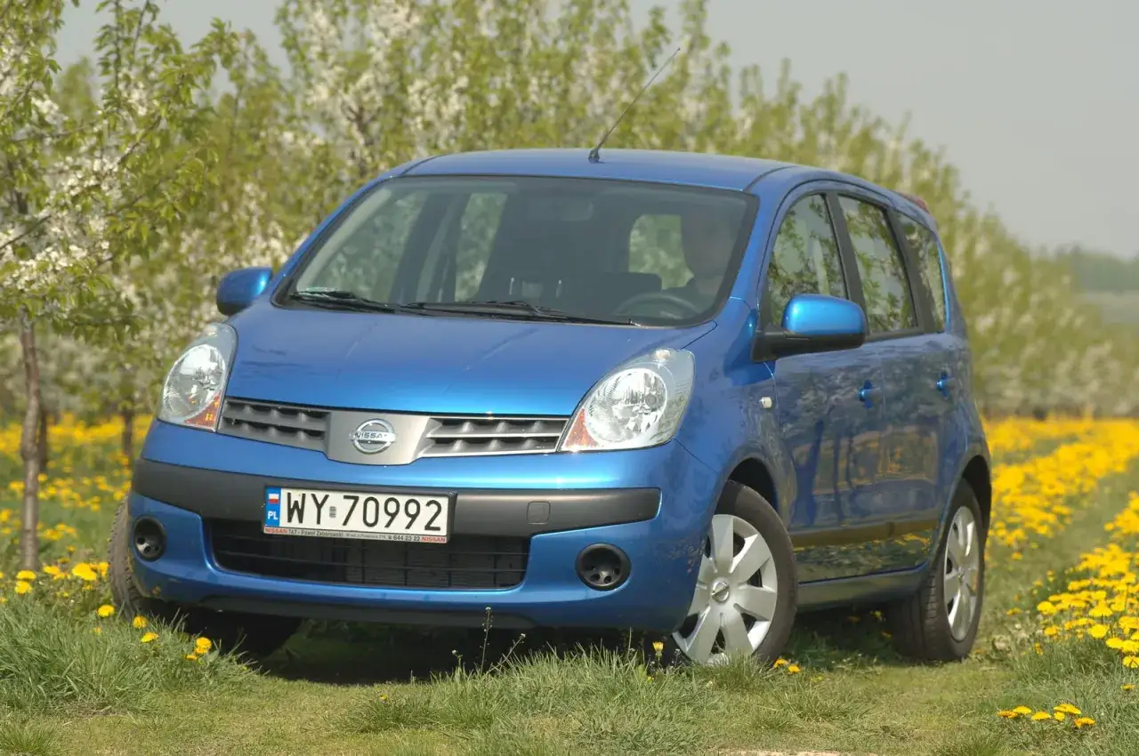 Niebieski Nissan Note 1.4, z widocznym kierowcą, stoi na łące pełnej żółtych kwiatów. Pojemność baku tego modelu jest idealna na wiosenne wycieczki.